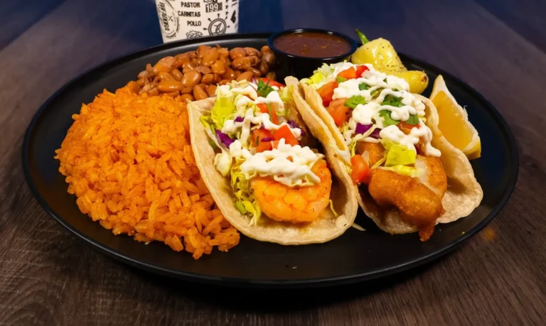 2 tacos on a plate topped with cabbage, pico de gallo, and cream, with rice, beans, and a fountain drink on the side.